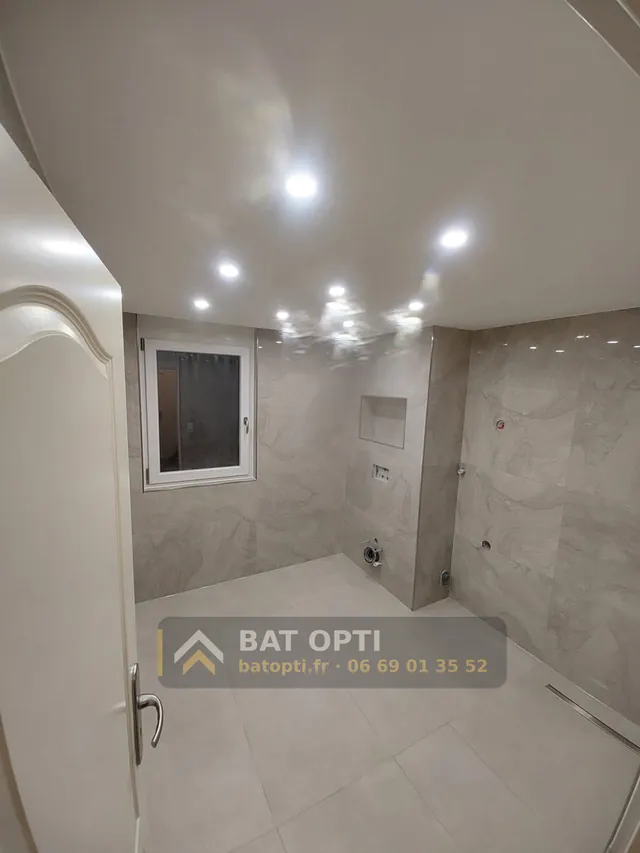 Salle de bain carrelée en grand format effet pierre, douche à l'italienne avec caniveau, niche murale et faux plafond à spots, sanitaires pas encore posés.